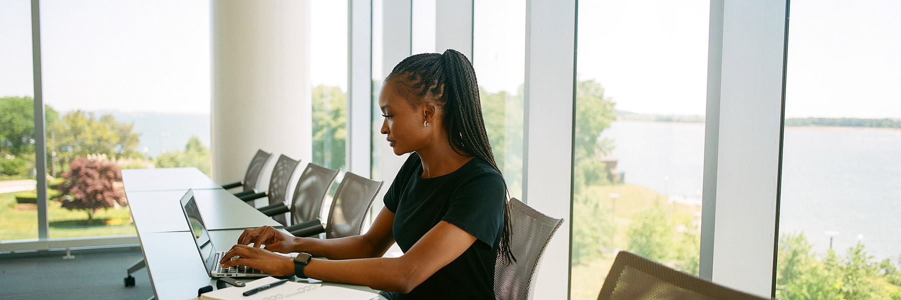 A UMass Boston student wearing a black dress sits at a table with her laptop in front of windows with a water view